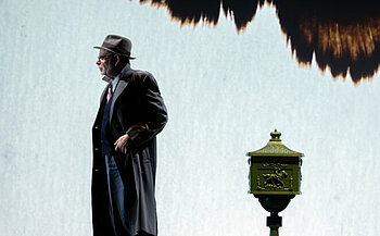 L’image montre un homme vêtu d’un manteau long noir, d’un costume trois pièces avec une cravate rouge, et d’un chapeau gris. Il porte également une barbe grise. Il se tient debout, légèrement tourné vers la gauche.  À sa droite (dans l'image), on aperçoit une boîte aux lettres ancienne de couleur verte avec des décorations dorées, posée sur un pied. L’arrière-plan est clair, avec une grande tache noire et brune en haut de l’image qui ressemble à une projection ou un motif abstrait. La scène est éclairée de façon à faire ressortir les silhouettes de l’homme et de la boîte aux lettres.