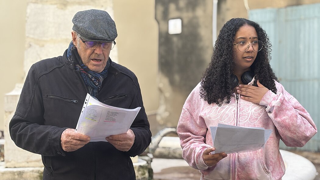     Requête réussie  Gemini said  L'image montre deux personnes, un homme plus âgé et une jeune femme, debout l'un à côté de l'autre à l'extérieur. Ils semblent être en train de lire ou de répéter quelque chose à partir de feuilles de papier qu'ils tiennent. Les Personnes      L'homme à gauche : Il porte une veste noire, un foulard à motifs bleus et bordeaux, une casquette grise et des lunettes. Il tient des feuilles de papier dans un protège-document transparent et semble lire attentivement à voix haute.      La femme à droite : Elle a de longs cheveux bouclés et porte un sweat à capuche rose délavé, un casque audio autour du cou et des lunettes à monture fine. Elle tient également des feuilles de papier et a une main posée sur sa poitrine, avec une expression qui suggère qu'elle chante ou qu'elle récite un texte avec émotion.  Le Décor      Ils se trouvent devant ce qui ressemble à une fontaine en pierre ou un muret circulaire.      L'arrière-plan est légèrement flou, montrant des murs de bâtiments aux tons neutres et une porte ou un volet bleu clair sur la droite.      L'ambiance générale suggère une répétition en plein air, peut-être pour une chorale, une pièce de théâtre ou un événement communautaire.