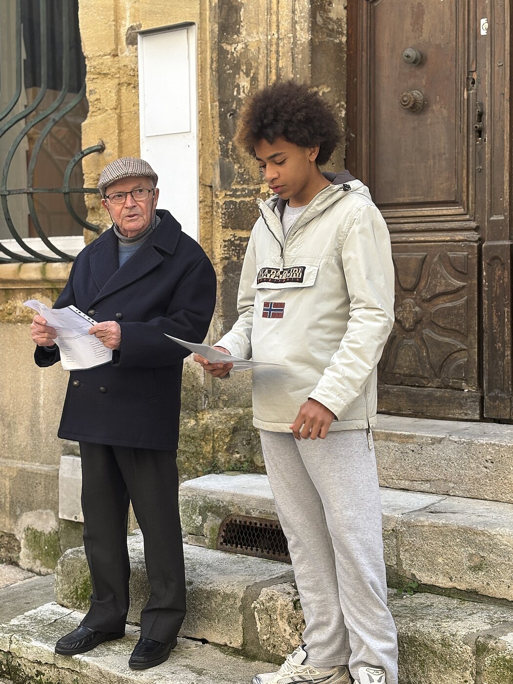 Cette image présente une scène de rue paisible et studieuse entre deux personnes d'âges différents, posant devant l'entrée d'un bâtiment ancien. Les Personnes      À gauche : Un homme plus âgé, portant des lunettes et une casquette en tweed. Il est vêtu élégamment d'un manteau caban bleu marine boutonné, d'un pantalon noir et de chaussures de ville sombres. Il tient une feuille de papier et semble expliquer ou lire quelque chose avec attention.      À droite : Un jeune homme aux cheveux volumineux et bouclés. Il porte une veste de type anorak claire (de la marque Napapijri) avec un pantalon de jogging gris et des baskets blanches. Il regarde également une feuille de papier qu'il tient à la main, semblant concentré sur son contenu.  Le Décor      L'entrée : Ils se tiennent sur des marches en pierre usées par le temps, devant une grande porte en bois sculpté de style classique.      L'architecture : Le mur est fait de pierres de taille de couleur ocre, typique de certaines régions européennes. On aperçoit sur la gauche une fenêtre protégée par une grille en fer forgé arrondie.      L'ambiance : La scène suggère un moment de transmission, de collaboration ou d'apprentissage, peut-être une répétition de texte ou la consultation de documents administratifs dans un cadre informel.