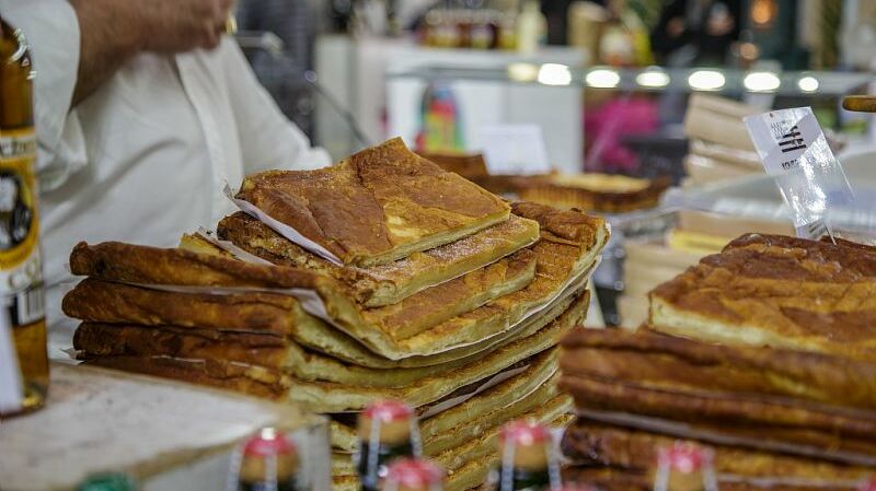 Cette image montre une pile de pains, probablement une spécialité locale, disposée sur une table. Les pains sont bien dorés et superposés, avec des couches bien définies, ce qui suggère qu'ils sont faits maison. En arrière-plan, on aperçoit des bouteilles de boisson et d'autres produits en vente, mais l'attention est principalement attirée par la pile de pains. Une personne en arrière-plan porte une chemise blanche, probablement un vendeur ou un artisan, mais elle est partiellement floue.