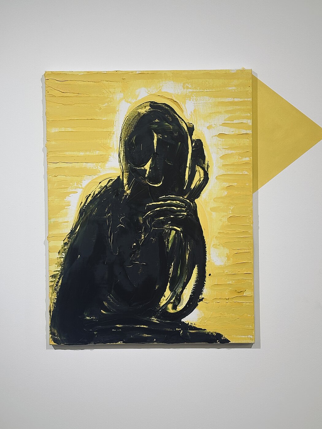 Cette œuvre est une peinture expressionniste saisissante, jouant sur un contraste binaire puissant et une texture très marquée. Voici une description détaillée de ses éléments :  ### Sujet et Composition  Au centre de la toile se détache une **silhouette sombre**, presque totalement noire, évoquant une figure humaine ou anthropomorphe.  * **La posture :** Le personnage semble assis ou recroquevillé, une main (ou une griffe) ramenée près du visage dans un geste qui peut suggérer la contemplation, la détresse ou le secret. * **Le visage :** Les traits sont effacés, laissant place à une forme abstraite et tourmentée. On devine des orbites ou des cavités, donnant à la figure un aspect spectral ou sculptural.  ### Palette de Couleurs  L'œuvre repose sur un dialogue entre deux teintes dominantes :  * **Le Jaune :** Un jaune vif, presque acide, occupe tout l'arrière-plan. Cette couleur apporte une luminosité agressive qui projette la figure noire vers l'avant. * **Le Noir :** Utilisé pour la figure centrale, il est dense et profond. Des touches de blanc et de jaune sont parfois entremêlées dans la silhouette pour suggérer du relief ou des reflets de lumière.  ### Technique et Texture  La matérialité de la peinture est un élément clé de l'œuvre :  * **Empâtements :** On observe des couches de peinture épaisses. À gauche et à droite du personnage, des stries horizontales régulières suggèrent l'utilisation d'un couteau à peindre ou d'un peigne à colle. * **Gestualité :** Le traitement de la silhouette est beaucoup plus libre et organique, avec des coups de pinceau larges et vigoureux qui contrastent avec la structure plus rigide du fond.  ### Éléments Contextuels  * **L'ombre géométrique :** Un triangle jaune plus foncé est projeté sur le mur blanc à droite du tableau. Cela crée un effet d'installation, brouillant la limite entre la toile et l'espace réel de la galerie. * **Ambiance :** L'ensemble dégage une sensation d'isolement et d'intensité psychologique. Le contraste violent évoque une forme de dualité entre la lumière (le fond) et l'obscurité (le sujet). 