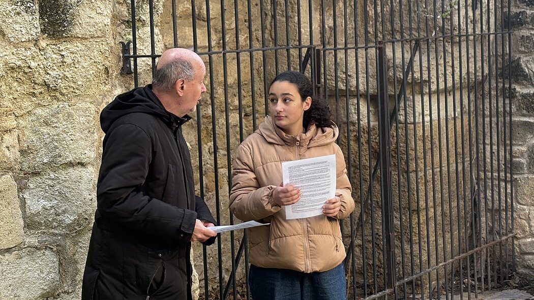 Cette image montre deux personnes, un homme et une jeune femme, se tenant devant une grille métallique noire et un mur en pierre ancienne.  ### Les Personnes  * **L'homme :** Situé à gauche, il est de profil. Il est chauve et porte une veste noire à capuche ainsi qu'un pantalon kaki. Il tient des feuilles de papier dans sa main gauche et semble s'adresser à la jeune femme. * **La jeune femme :** Située à droite, elle regarde vers l'homme. Elle a des cheveux foncés attachés et porte une doudoune beige (ou marron clair) avec un jean large foncé. Elle tient également une feuille de papier avec du texte imprimé.  ### Le Décor  * **Arrière-plan :** Ils se tiennent devant une **grille haute en fer forgé** composée de barreaux verticaux. Derrière cette grille, on aperçoit une structure en pierre, suggérant peut-être l'entrée d'un monument historique ou d'un site ancien. * **Texture :** Sur la gauche, le mur est fait de **pierres irrégulières** aux tons clairs et terreux. Le sol semble être un pavage ou un chemin en terre battue.  ### L'Atmosphère  L'interaction semble sérieuse ou informative....