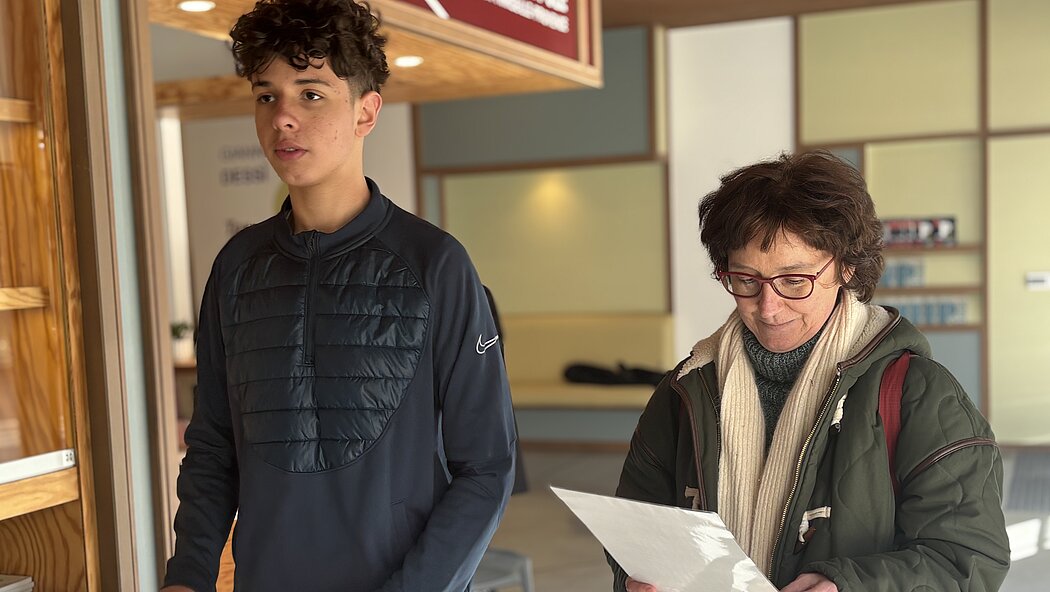     Requête réussie  Gemini said  Cette image montre deux personnes, un jeune homme et une femme plus âgée, se tenant debout à l'intérieur de ce qui semble être un lieu public ou un centre d'information.  Voici une description détaillée de la scène : Les Personnes      Le jeune homme : Il se tient sur la gauche, regardant vers l'avant avec une expression neutre. Il a des cheveux bruns bouclés et porte un haut de sport bleu marine à manches longues avec une partie matelassée sur le torse et un logo Nike sur le bras.      La femme : Elle se tient à droite, légèrement en retrait, et regarde attentivement un document ou une feuille de papier blanc qu'elle tient à deux mains. Elle porte des lunettes à monture rouge, une écharpe beige, un pull à col roulé gris et une veste d'hiver vert olive.  L'Environnement      Le décor : L'arrière-plan suggère un bâtiment moderne avec des menuiseries en bois clair. On aperçoit une étagère ou un présentoir à gauche contenant des brochures ou des dépliants.      La signalisation : Au-dessus d'eux, une partie d'un panneau rouge est visible, comportant du texte blanc dont le mot « METROPOLE ».      L'ambiance : La lumière semble naturelle et provient de la droite, créant des ombres douces et une atmosphère calme.