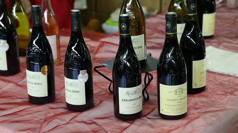 Cette image montre plusieurs bouteilles de vin disposées sur une table. Les bouteilles sont alignées sur une nappe rouge. On peut distinguer des bouteilles avec des étiquettes visibles, indiquant des appellations telles que Château-Neuf-du-Pape et Côtes du Rhône. Certaines bouteilles sont posées sur un support métallique pour les maintenir debout. En arrière-plan, on peut voir un peu de flou, suggérant une autre activité ou un autre espace.
