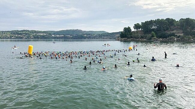 L'image montre un groupe de nageurs participant à une compétition ou un événement aquatique dans un plan d'eau. Les participants, qui portent des bonnets de bain de différentes couleurs, sont répartis sur la surface de l'eau. Certains sont en train de nager, tandis que d'autres semblent marcher ou se tenir debout dans l'eau. Un dispositif flottant de couleur jaune, probablement un repère de course, est visible au centre de l'image. En arrière-plan, on aperçoit des collines boisées et quelques personnes sur des kayaks ou des planches, surveillant l'événement. Le ciel est partiellement nuageux.
