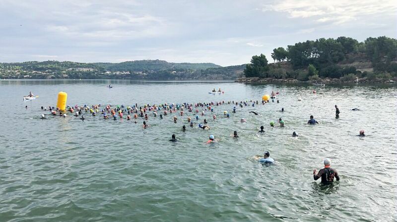 L'image montre un groupe de nageurs participant à une compétition ou un événement aquatique dans un plan d'eau. Les participants, qui portent des bonnets de bain de différentes couleurs, sont répartis sur la surface de l'eau. Certains sont en train de nager, tandis que d'autres semblent marcher ou se tenir debout dans l'eau. Un dispositif flottant de couleur jaune, probablement un repère de course, est visible au centre de l'image. En arrière-plan, on aperçoit des collines boisées et quelques personnes sur des kayaks ou des planches, surveillant l'événement. Le ciel est partiellement nuageux.