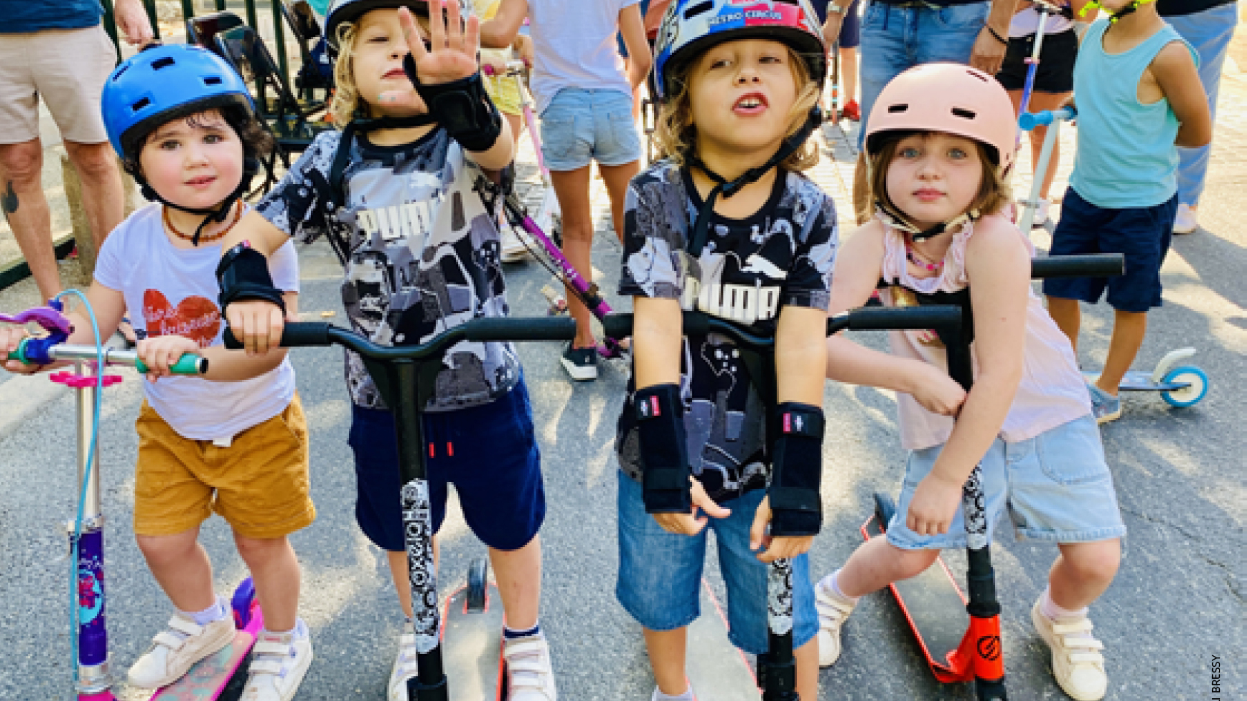 On voit un groupe d’enfants, principalement au premier plan, qui font de la trottinette dans un cadre urbain ou lors d’un événement organisé. Ils portent tous des casques de protection (bleu, rouge, rose) et certains ont des gants ou protections aux poignets. Il y a quatre enfants au centre, alignés côte à côte, posant pour la photo. En arrière-plan, d'autres enfants et des adultes semblent attendre ou assister à la scène.  Les enfants ont une attitude détendue, joyeuse, et certains font des gestes (comme un signe de la main). Les vêtements sont estivaux, ce qui suggère une journée ensoleillée et festive.