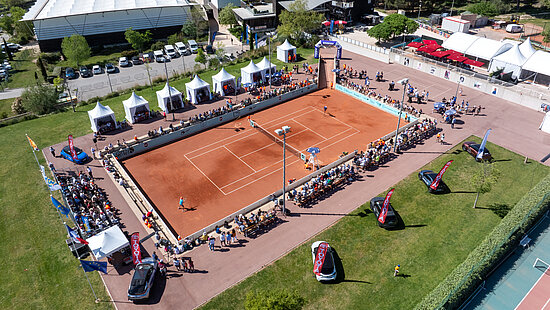 Il s’agit d’une vue aérienne en plongée (prise par un drone) montrant le court central de tennis d'Istres lors d'un tournoi international. L'image dégage une impression de clarté, de dynamisme et d'organisation, sous un soleil printanier éclatant. Le court de tennis : le point focal  Au centre de l'image se trouve un court de tennis en terre battue d'un orange brique très vif, qui contraste avec le vert environnant.      On y voit deux joueurs en plein échange (de minuscules silhouettes en mouvement).      Le court est impeccablement tracé de lignes blanches.      Des tribunes basses et temporaires entourent les quatre côtés du terrain, remplies de spectateurs assis qui forment une bordure colorée tout autour du jeu.