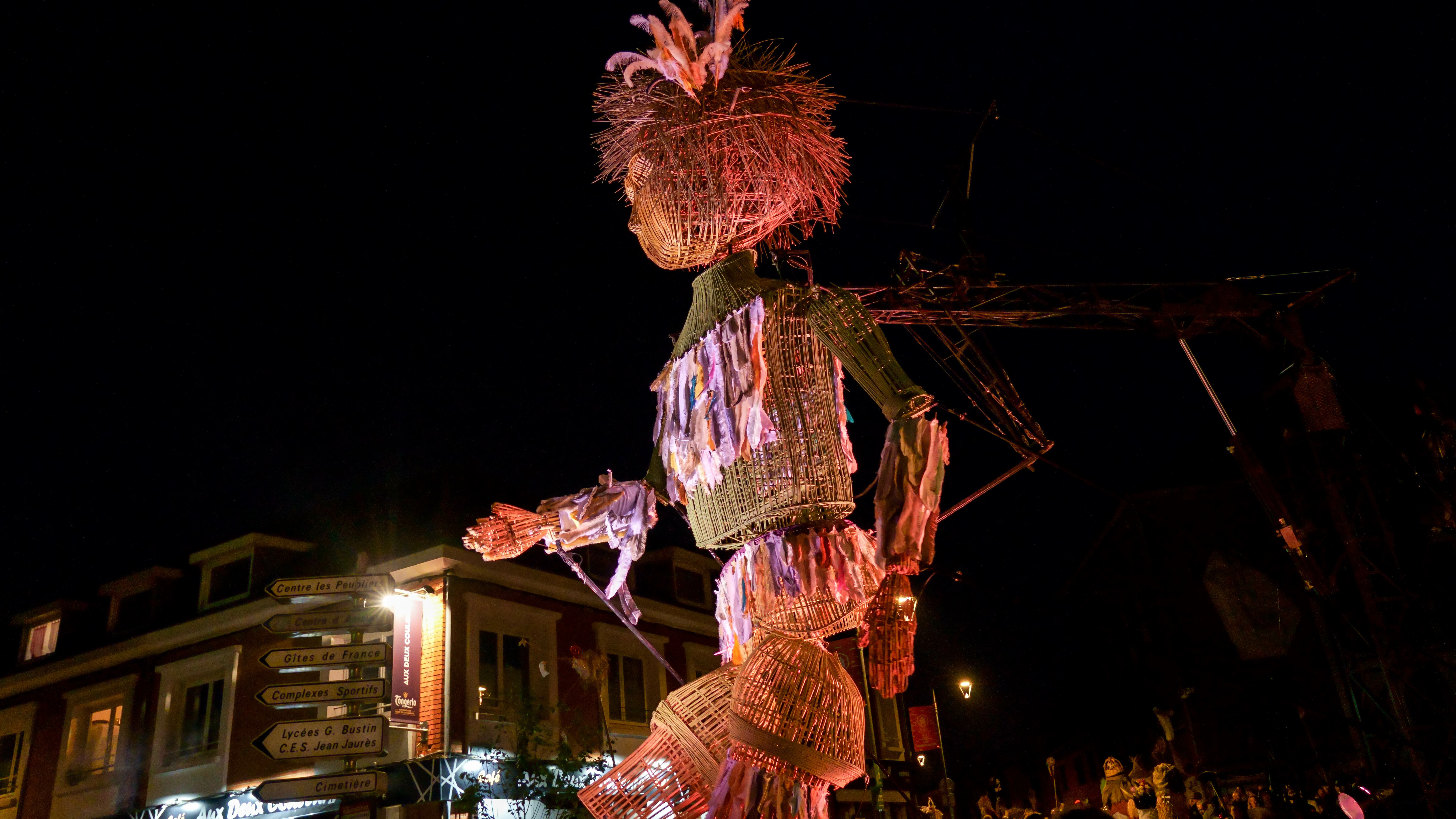 photo du spectacle la cabane à plumes L'image montre deux structures illuminées faites de matériaux tressés ou entrelacés, probablement en fibres végétales. La structure de gauche représente une silhouette humaine de profil, tournée vers la droite. Elle tient dans sa main un objet ressemblant à un bâton ou perchoir, sur lequel se trouve une autre structure plus petite, évoquant la forme d’un oiseau. Les deux structures sont éclairées de l’intérieur, produisant des teintes chaudes (jaune, orange) avec quelques reflets bleutés sur le bas du perchoir. L’arrière-plan est entièrement noir, mettant en valeur les formes lumineuses. Une signature au bas de l’image indique « © Sébastien CARLIER / PNRLG ».