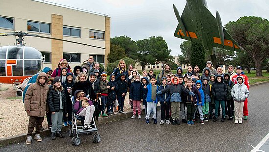 Cette photo montre un grand groupe composé d'enfants et de quelques adultes. Ils posent devant des aéronefs (un hélicoptère et un avion de chasse) lors de la visite de la BA125