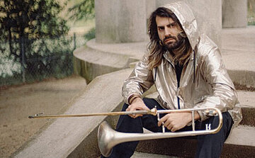 Sur cette image, un homme est assis sur des marches en pierre, à l’extérieur, tenant un trombone. Il porte une veste à capuche argentée, qui reflète la lumière, ainsi qu’un pantalon sombre. Ses cheveux sont longs et ondulés, et il a une barbe fournie. La capuche est relevée sur sa tête.  L’homme regarde l’objectif avec une expression neutre. En arrière-plan, on aperçoit une structure en pierre cylindrique et, plus loin, une clôture métallique et des arbres, suggérant un parc ou un jardin public. L’atmosphère générale est calme et naturelle.