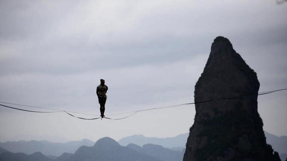 Funambule Pablo Signoret  -  ChatGPT a dit :  L’image montre une personne debout sur une corde tendue en hauteur entre deux points. La personne porte un casque et un harnais de sécurité attaché à la corde. À l’arrière-plan, on voit une grande formation rocheuse en forme de pic ainsi que des montagnes lointaines sous un ciel nuageux. L’ensemble de la scène est photographié avec une faible luminosité ambiante, donnant un ton gris-bleu à l'image.