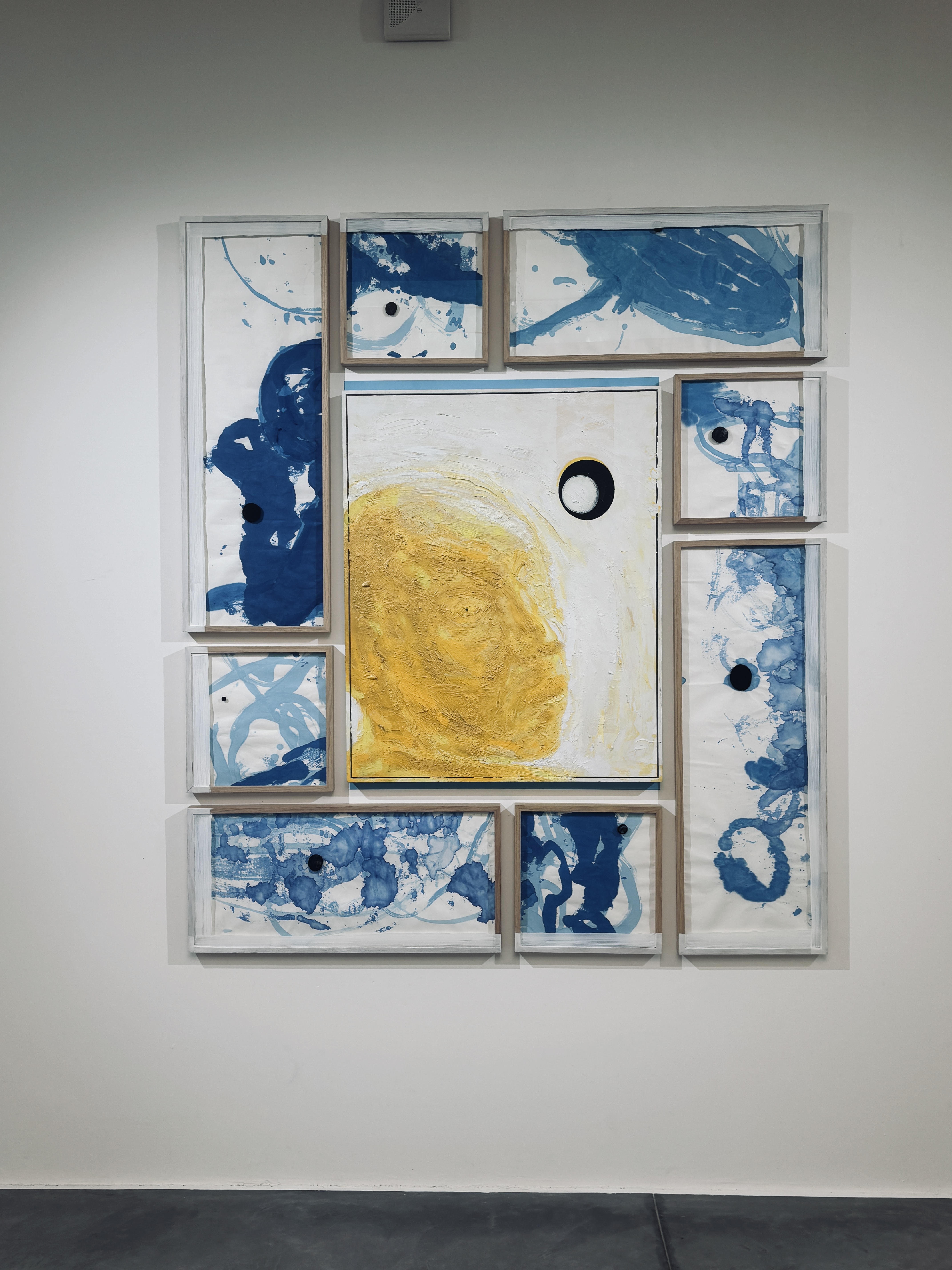 Cette œuvre est une composition murale multi-panneaux qui allie peinture abstraite et assemblage géométrique. Elle se compose d'un panneau central entouré de huit cadres rectangulaires de tailles variées, créant une structure globale équilibrée mais dynamique.  Voici une description détaillée de ses composants :  ### Le Panneau Central  * **Contenu :** Il présente une forme organique jaune ocre, texturée et dense, qui occupe la majeure partie de la surface blanche. On y voit également un cercle noir parfait, entouré d'un fin liseré blanc et noir, rappelant une éclipse ou un œil. * **Texture :** La peinture jaune semble appliquée en couches épaisses, créant un relief qui contraste avec la planéité des éléments environnants.  ### Les Cadres Périphériques  * **Style :** Huit cadres en bois clair entourent le centre. Ils contiennent des compositions à l'encre ou à l'aquarelle bleue sur fond blanc. * **Motifs :** Le bleu est appliqué de manière fluide, avec des effets d'éclaboussures, de taches et de lignes sinueuses évoquant des formes aquatiques ou cellulaires. * **Points Noirs :** Presque chaque panneau satellite contient un petit disque noir plein, créant un rappel visuel avec le cercle du panneau central et guidant l'œil à travers toute la structure.  ### Composition et Matériaux  * **Palette :** L'œuvre repose sur un contraste primaire entre le **jaune solaire**, le **bleu profond** et des touches de **noir**, le tout sur un fond blanc éclatant. * **Structure :** L'agencement des cadres rappelle une grille déconstruite. L'espace vide entre les cadres fait partie intégrante de l'œuvre, laissant respirer chaque élément.  L'ensemble dégage une impression de cosmologie ou de cartographie abstraite, où des éléments micro (les taches d'encre) et macro (la forme jaune centrale) coexistent. 