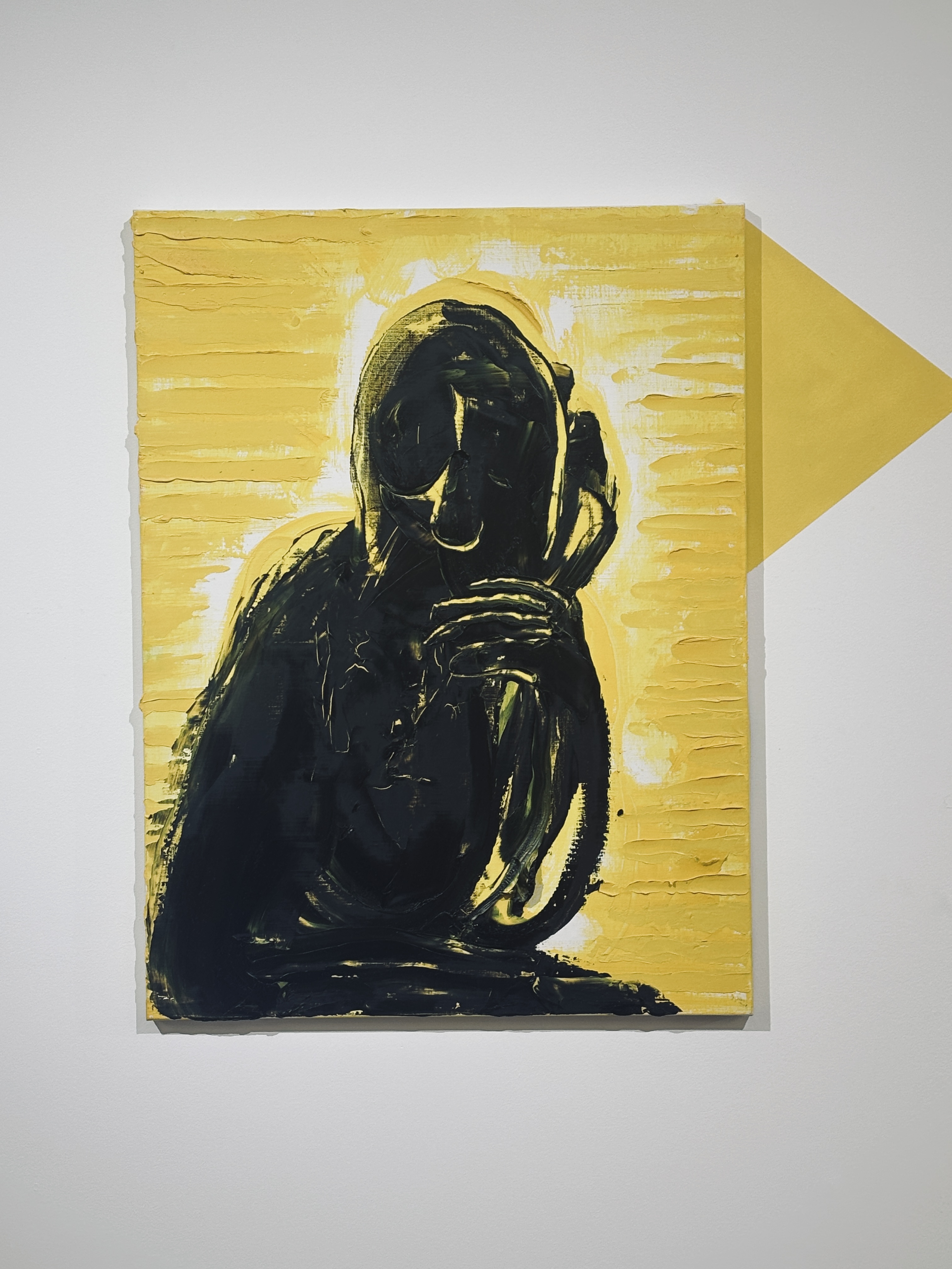 Cette œuvre est une peinture expressionniste saisissante, jouant sur un contraste binaire puissant et une texture très marquée. Voici une description détaillée de ses éléments :  ### Sujet et Composition  Au centre de la toile se détache une **silhouette sombre**, presque totalement noire, évoquant une figure humaine ou anthropomorphe.  * **La posture :** Le personnage semble assis ou recroquevillé, une main (ou une griffe) ramenée près du visage dans un geste qui peut suggérer la contemplation, la détresse ou le secret. * **Le visage :** Les traits sont effacés, laissant place à une forme abstraite et tourmentée. On devine des orbites ou des cavités, donnant à la figure un aspect spectral ou sculptural.  ### Palette de Couleurs  L'œuvre repose sur un dialogue entre deux teintes dominantes :  * **Le Jaune :** Un jaune vif, presque acide, occupe tout l'arrière-plan. Cette couleur apporte une luminosité agressive qui projette la figure noire vers l'avant. * **Le Noir :** Utilisé pour la figure centrale, il est dense et profond. Des touches de blanc et de jaune sont parfois entremêlées dans la silhouette pour suggérer du relief ou des reflets de lumière.  ### Technique et Texture  La matérialité de la peinture est un élément clé de l'œuvre :  * **Empâtements :** On observe des couches de peinture épaisses. À gauche et à droite du personnage, des stries horizontales régulières suggèrent l'utilisation d'un couteau à peindre ou d'un peigne à colle. * **Gestualité :** Le traitement de la silhouette est beaucoup plus libre et organique, avec des coups de pinceau larges et vigoureux qui contrastent avec la structure plus rigide du fond.  ### Éléments Contextuels  * **L'ombre géométrique :** Un triangle jaune plus foncé est projeté sur le mur blanc à droite du tableau. Cela crée un effet d'installation, brouillant la limite entre la toile et l'espace réel de la galerie. * **Ambiance :** L'ensemble dégage une sensation d'isolement et d'intensité psychologique. Le contraste violent évoque une forme de dualité entre la lumière (le fond) et l'obscurité (le sujet). 