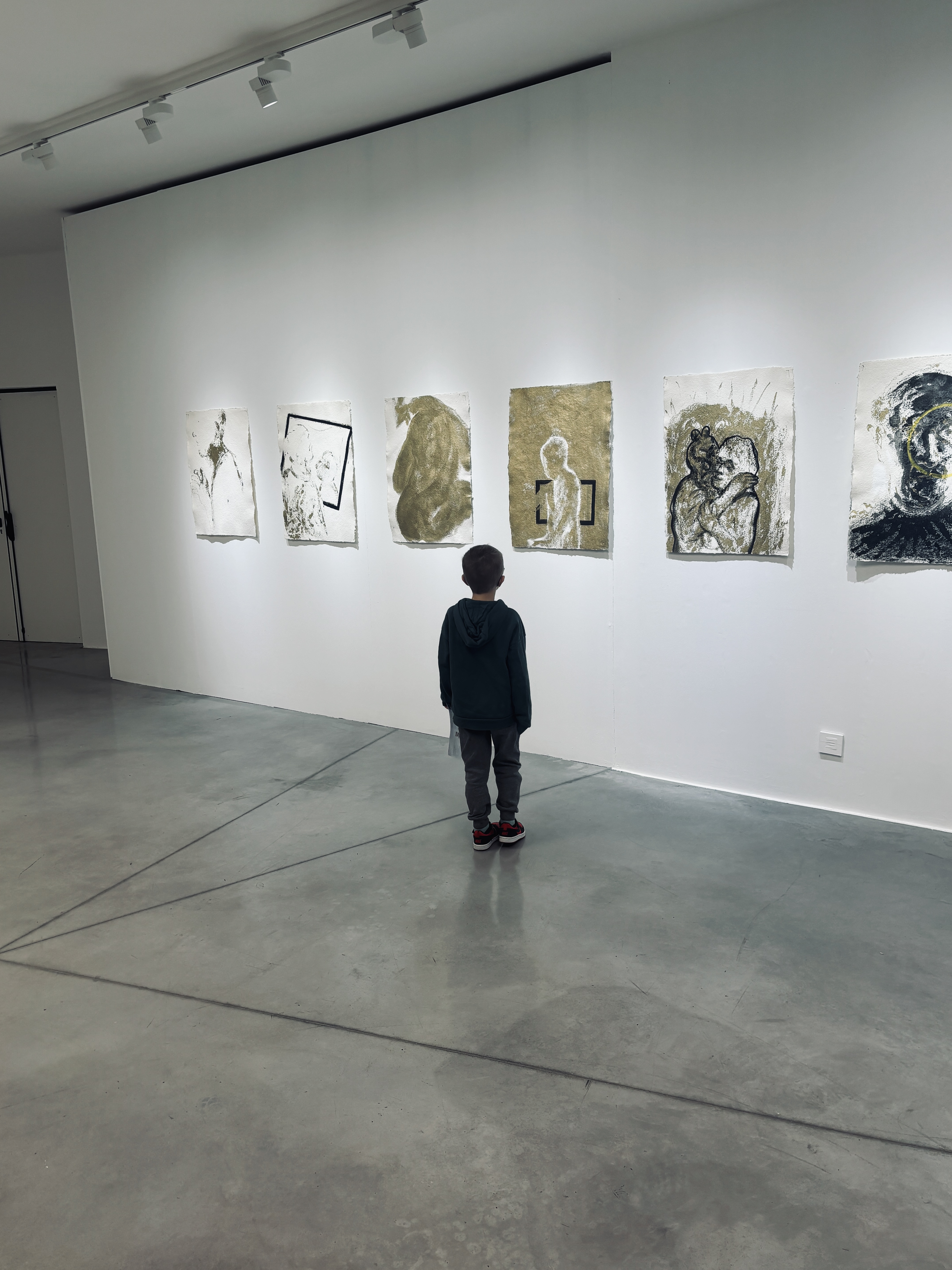 Cette image présente une scène paisible et contemplative dans une **galerie d'art moderne** ou un musée. Voici une description détaillée de la composition :  ### Le sujet principal  Au centre de l'image, un **jeune enfant** est vu de dos. Il porte un sweat à capuche foncé, un pantalon gris et des baskets noires avec des semelles rouges. Sa posture immobile suggère qu'il est absorbé par l'observation des œuvres devant lui, créant un contraste frappant entre la petite taille de l'enfant et l'imposante rangée de tableaux.  ---  ### L'exposition artistique  Sur un mur blanc épuré, six œuvres d'art sont alignées horizontalement. Elles semblent être des techniques mixtes sur papier à bords irréguliers :  * **Style :** Les œuvres utilisent une palette minimaliste composée de noir, de blanc et de tons dorés ou ocres. * **Contenu :** On y distingue des formes abstraites, des silhouettes humaines et des motifs géométriques (comme des cadres noirs dessinés à l'intérieur des compositions). * **Éclairage :** Des projecteurs fixés au plafond (visibles en haut à gauche) dirigent une lumière douce sur chaque pièce, accentuant les textures du papier.  ---  ### L'environnement et l'ambiance  * **L'espace :** La pièce est vaste et minimaliste. Le sol est en **béton poli gris**, reflétant subtilement la silhouette de l'enfant et la lumière de la pièce. * **Lignes de force :** Des lignes de jointure au sol convergent vers le bas de l'image, renforçant la perspective et guidant le regard vers l'enfant. * **Atmosphère :** L'ensemble dégage une impression de calme, de curiosité et de respect pour l'art. Le dépouillement de la salle met l'accent sur l'interaction silencieuse entre le spectateur et l'œuvre. 