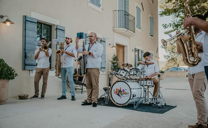 L'image montre un groupe de musiciens jouant à l'extérieur devant un bâtiment beige avec des volets bleus. Il y a six hommes visibles, tous vêtus de chemises blanches et de pantalons clairs. Un batteur est assis à une batterie blanche avec le nom "Blaye's" inscrit sur la grosse caisse. Les autres musiciens jouent des instruments à vent, notamment un trombone, un saxophone baryton, un saxophone alto et deux trompettes. Le sol est en béton clair et il y a quelques plantes en pot près des murs. Le bâtiment a plusieurs fenêtres, une petite porte en bois, un balcon en fer forgé, et des volets ouverts. Il fait jour et le ciel est dégagé.