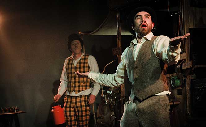 L'image montre deux hommes en costumes, sur scène dans un décor sombre. Le premier homme porte un gilet gris clair, une chemise blanche et un chapeau, et semble s'exprimer avec enthousiasme, les bras écartés. Le deuxième homme, en arrière-plan, porte un costume à carreaux jaunes et oranges, avec un chapeau. Il tient un objet rouge, possiblement une sorte de récipient. L'éclairage est tamisé, mettant en valeur les personnages et créant une ambiance théâtrale. Ils semblent faire partie d'une performance ou d'une pièce de théâtre.