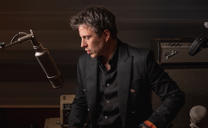 Photo de l'artiste Charles Pasi - L’image montre un homme aux cheveux courts et ébouriffés, vêtu d'une veste noire et d'une chemise sombre. Il est penché légèrement en avant, regardant vers le bas à sa droite. Devant lui se trouve un grand microphone de style vintage. À l’arrière-plan, on aperçoit un amplificateur de guitare de marque Fender et d'autres équipements audio, suggérant un environnement de studio d'enregistrement. L’éclairage est tamisé, avec un fond sombre.