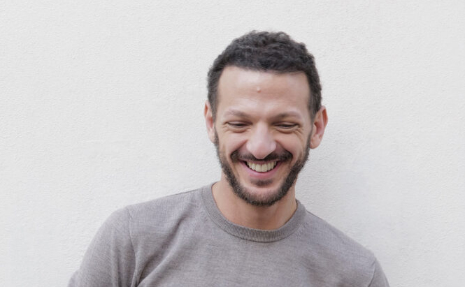 L’image montre une l'acteur Vincent DEDIENNE debout devant un mur blanc uni. Elle porte un pull léger de couleur gris clair. Elle a les cheveux courts et une barbe. Elle sourit en regardant vers le bas ou les yeux fermés. La photo est prise en lumière naturelle, avec un cadrage centré sur le haut du corps.