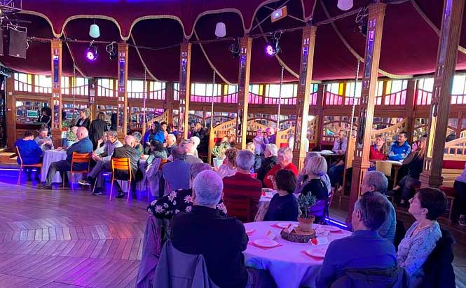 L'image montre un groupe de personnes assises à des tables rondes dans un espace intérieur. Les invités semblent être principalement des personnes âgées. L'environnement est un lieu décoré avec des murs en bois et des fenêtres colorées, créant une ambiance chaleureuse et conviviale. La lumière est tamisée, avec des spots de couleur violette qui ajoutent une touche festive. Certains invités regardent vers le devant, tandis que d'autres semblent discuter entre eux. On peut voir des décorations sur les tables, comme des plantes et des assiettes. Le lieu semble être un espace pour une réception ou un événement spécial.