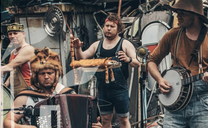 Photo du groupe Steve N Seagulls - L’image montre un groupe de quatre hommes en train de jouer de la musique dans un décor extérieur encombré d’objets divers. Un homme à gauche porte une couronne dorée et tient une baguette. Un autre homme, au centre, tient une marionnette en forme de saucisse. Un troisième homme, à l'avant, joue de l’accordéon et porte une coiffe en forme de tête de lion. Le quatrième homme, à droite, joue du banjo et porte un chapeau à larges bords ainsi que des bretelles. Les vêtements sont dépareillés et de style décalé. Un microphone ancien est visible au centre.