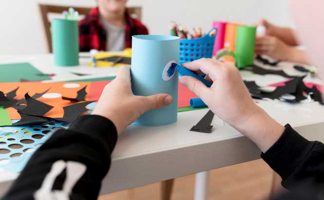 L'image montre un atelier de bricolage avec des enfants en arrière-plan. Un enfant est concentré sur l'assemblage d'un objet à partir d'un tube de papier bleu. L'enfant tient un œil en plastique et l'applique sur le tube à l'aide d'un bâton de colle. Sur la table, on peut voir plusieurs feuilles de papier de couleurs vives (orange, vert, bleu), des formes découpées en noir et en blanc, ainsi que des ciseaux et des outils de collage. Des crayons et des fournitures sont rangés dans un panier coloré à proximité, créant un environnement animé et créatif pour l'activité manuelle.
