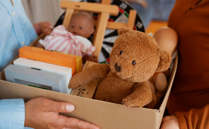 Une peluche en forme d’ours brun, bien visible au premier plan.  Une poupée habillée d’une tenue rose et blanche.  Un jeu de fléchettes en arrière-plan.  Plusieurs cadres ou livres empilés sur le côté gauche de la boîte.  Un petit chevalet en bois.  D’autres objets difficilement identifiables, partiellement cachés.