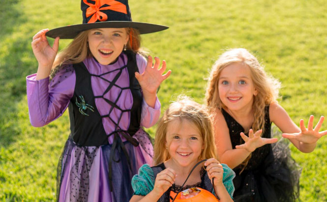 À gauche, un enfant porte un grand chapeau noir décoré d’un ruban orange et une robe violette avec un gilet noir.  Au centre, un autre enfant sourit en tenant un seau orange en forme de citrouille.  À droite, un enfant porte une robe noire sans manches et a les bras levés vers l’avant.  Ils se trouvent dehors, sur une pelouse verte, et semblent posés côte à côte.