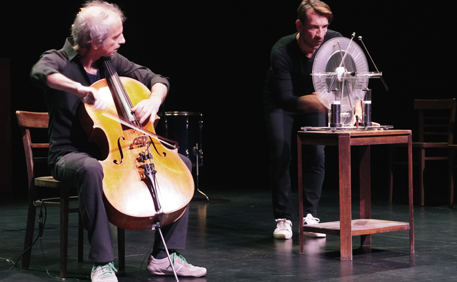 Sur cette image, deux hommes se tiennent sur une scène noire. À gauche, un homme aux cheveux gris joue du violoncelle, assis sur une chaise en bois. Il porte des vêtements sombres et des baskets vert clair et blanches. L’instrument repose entre ses jambes, et il utilise un archet pour jouer. À droite, un autre homme interagit avec un dispositif mécanique posé sur une petite table en bois. Ce dispositif comprend une roue transparente montée verticalement, des tiges et plusieurs objets techniques ou scientifiques. Il semble en pleine manipulation de l'appareil. L'homme est également vêtu de noir et porte des baskets blanches. L’arrière-plan est sombre, et plusieurs chaises en bois sont visibles, suggérant un espace scénique ou de performance.