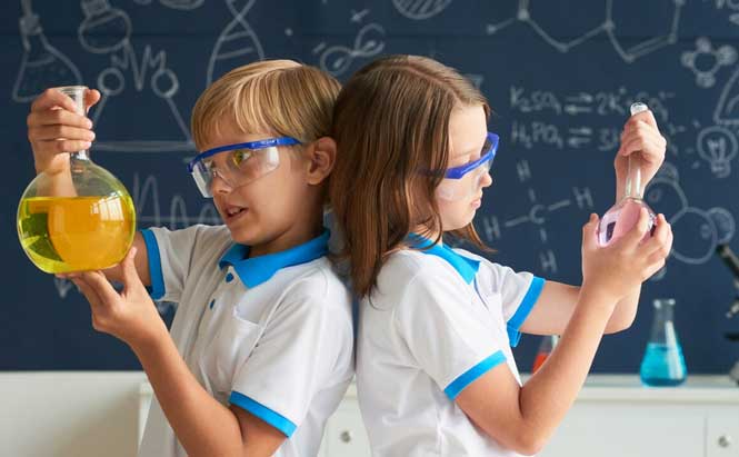 Sur l’image, on voit deux enfants vêtus de polos blancs avec des liserés bleus. Ils portent des lunettes de protection transparentes et bleues.  L’enfant à gauche tient un grand ballon de verre contenant un liquide jaune.  L’enfant à droite tient un ballon de verre plus petit contenant un liquide rose.  Ils se tiennent dos à dos.  En arrière-plan, il y a un tableau bleu foncé avec des dessins et formules scientifiques tracés à la craie blanche.  Sur une table derrière eux, on distingue d’autres récipients de laboratoire, dont un erlenmeyer contenant un liquide bleu.