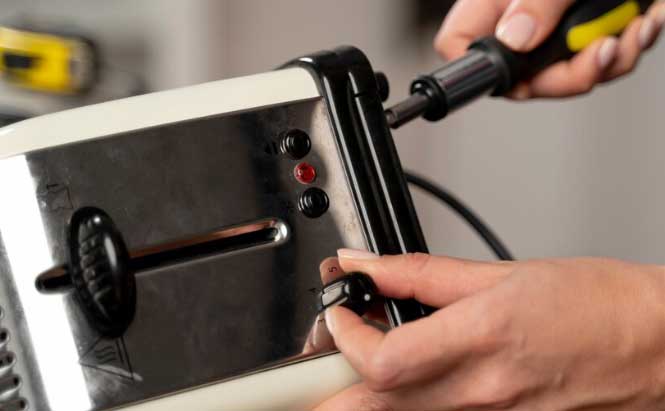 L’image montre un appareil électroménager de couleur blanche et métallique. Une main tient un tournevis inséré dans une vis située en haut de l’appareil. Une autre main tient une pièce noire fixée sur le côté. Sur la façade de l’appareil, il y a un levier noir, une fente horizontale et trois boutons ronds noirs avec un petit voyant rouge.