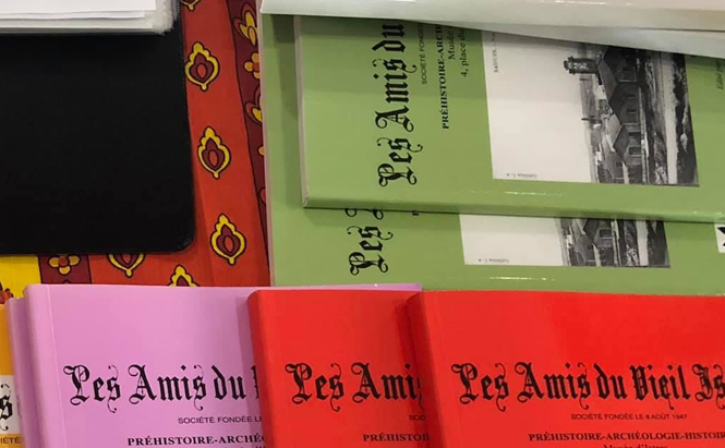 L’image montre plusieurs publications empilées sur une surface recouverte d’un tissu rouge à motifs jaunes et noirs. Les couvertures des publications sont de différentes couleurs : vert clair, rouge, rose et une partie jaune visible en haut à gauche. Le titre commun visible sur plusieurs couvertures est « Les Amis du Vieil », suivi d’un texte en plus petit qui mentionne « PRÉHISTOIRE – ARCHÉOLOGIE – HISTOIRE ».  Les titres sont imprimés en lettres noires avec une police de style ancien. Deux des couvertures vertes comportent aussi une photo en noir et blanc représentant des bâtiments.  En haut à gauche, il y a une pile de feuilles blanches posées sur un objet noir rectangulaire.