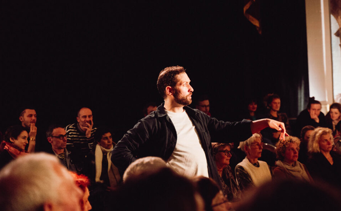 L’image montre un homme debout au centre d’un espace entouré de personnes assises. Il porte un t-shirt blanc et une veste foncée. Son bras droit est tendu vers le côté, et il regarde dans cette direction. Autour de lui, un public nombreux observe attentivement. L’éclairage est chaud, concentré principalement sur l’homme au centre, tandis que l’arrière-plan est sombre. Les spectateurs sont habillés de manière variée, assis en rangées proches les unes des autres.