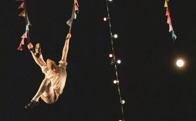 L’image montre une personne suspendue dans les airs, accrochée à deux cordes ornées de petits fanions colorés. Elle porte des vêtements clairs et des chaussures noires. À côté des cordes, une guirlande lumineuse est également suspendue, avec des ampoules allumées de différentes couleurs. Le ciel est sombre, et une pleine lune est visible en arrière-plan.