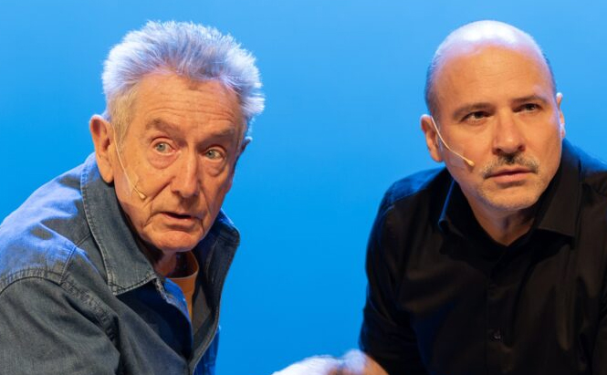 Photo des acteurs Christian Ubl et Gilles Clément  - Deux hommes sont visibles sur l’image, tous deux équipés de micros serre-tête. Celui de gauche a les cheveux gris en bataille, porte une chemise en jean et regarde légèrement vers la gauche avec une expression surprise. Celui de droite est chauve, a une moustache et porte une chemise noire. Il regarde droit devant lui avec un air sérieux. L’arrière-plan est uniformément bleu.