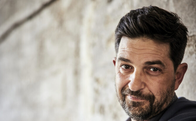 Photo de Tiago Rodrigues Un homme est visible sur l’image. Il a les cheveux foncés, courts et coiffés vers l’arrière, ainsi qu’une barbe fournie. Il regarde l’objectif avec un léger sourire. L’arrière-plan est un mur en pierre clair, flou, mettant en valeur le sujet en premier plan. La lumière est douce et naturelle.