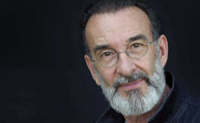 Photo de l'acteur Robin Renucci L’image montre un homme âgé avec une barbe grise bien taillée et des cheveux foncés en arrière. Il porte des lunettes rondes à monture fine. Il est vêtu d’un vêtement sombre, probablement une veste en jean ou en tissu similaire. L’arrière-plan est uni et noir, mettant l’accent sur son visage. Il regarde directement vers l’objectif avec une expression douce.
