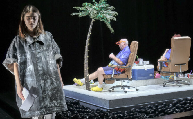 L’image montre trois personnes sur une scène. Au premier plan, une femme debout porte un vêtement ample de couleur grise et tient des feuilles de papier. À l’arrière-plan, deux personnes sont assises sur des chaises de bureau marron, sur une plateforme surélevée. Elles portent des vêtements violets et des chaussures jaunes. Une fausse palme éclairée est plantée au centre de la plateforme, à côté d’une glacière bleue et d’objets divers. Le sol autour de la plateforme est recouvert de nombreuses boules noires. L’arrière-plan est sombre.