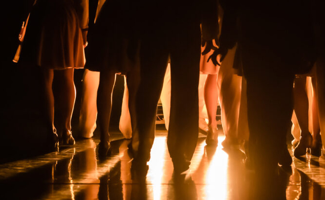 L’image montre un groupe de personnes debout, vues de dos et principalement cadrées au niveau des jambes. Elles sont éclairées par une forte lumière dorée provenant du sol ou de l’arrière-plan, créant des ombres marquées et des reflets sur une surface brillante. Les tenues varient entre jupes et pantalons. L’ambiance générale est sombre, contrastée par l’éclairage intense.