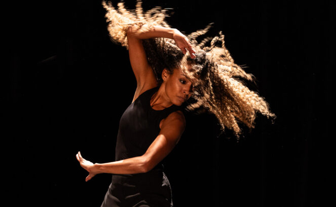 photo de la danseuse Ana Pérez L’image montre une personne aux cheveux longs et bouclés en mouvement, portant un débardeur noir et un pantalon foncé. Elle est photographiée en plein geste de danse, un bras levé au-dessus de la tête et l'autre dirigé vers le bas. Ses cheveux sont projetés en l’air, suggérant un mouvement énergique. L’arrière-plan est sombre, mettant en valeur la silhouette et le mouvement du sujet.