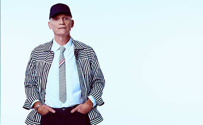 Photo de Gaetan Roussel On voit une personne photographiée en pied jusqu’au haut des cuisses, debout face à l’appareil. Elle porte une casquette noire.  Elle est vêtue d’une chemise blanche, d’une cravate claire avec de fines bandes colorées, et d’une veste à manches longues à rayures verticales noires et blanches.  Le pantalon est foncé. Les mains sont placées dans les poches du pantalon.  Le visage est visible, sans barbe. Le regard est dirigé vers l’avant.  Le fond de l’image est uni et très clair, proche du blanc ou du bleu très pâle, sans élément visible. L’éclairage est uniforme, sans ombre marquée.