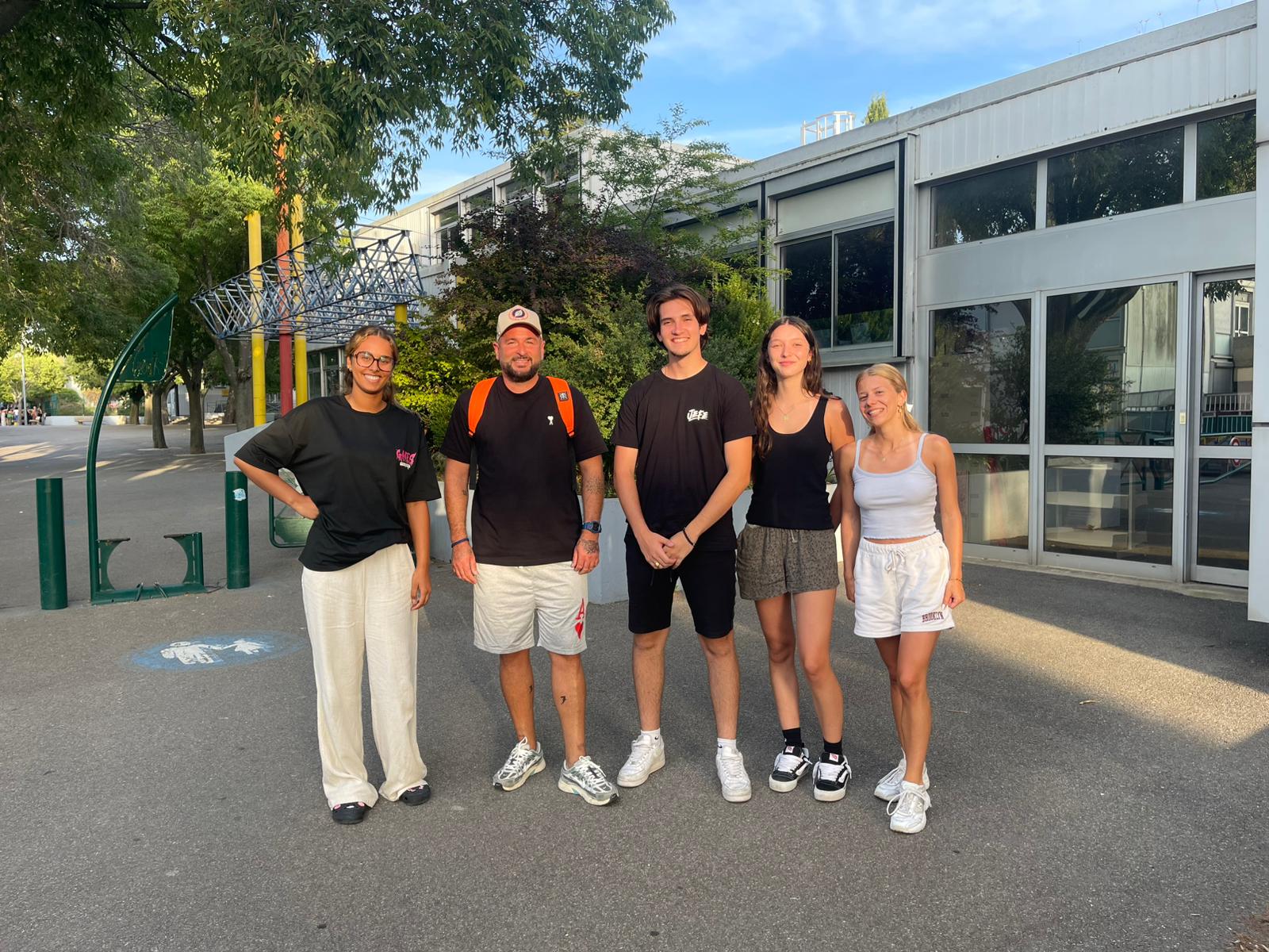L'équipe d'animation en photo Sur la photo, cinq personnes sont debout à l’extérieur sur une zone goudronnée, devant un bâtiment moderne avec de grandes fenêtres. Elles posent pour la photo, souriantes, en rang côte à côte.  De gauche à droite :  La première personne porte un t-shirt noir et un pantalon ample beige, avec des chaussures noires.  La deuxième personne porte un t-shirt noir, un short clair, une casquette beige et un sac à dos orange.  La troisième personne porte un t-shirt noir et un short noir, avec des chaussures blanches.  La quatrième personne porte un débardeur noir et un short gris, avec des chaussures noires et blanches.  La cinquième personne porte un débardeur blanc et un short blanc, avec des baskets blanches.  En arrière-plan, on voit des arbres, une structure métallique colorée (avec des poteaux jaunes, rouges et violets), et un bâtiment aux murs clairs et aux grandes baies vitrées. Il fait beau et le ciel est bleu.
