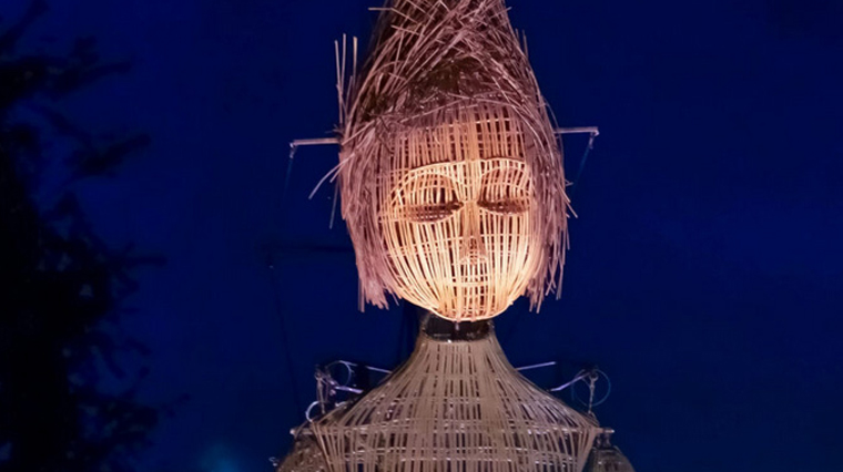 L’image montre une grande structure représentant une silhouette humaine stylisée, construite en matériaux entrelacés de type osier ou bois. La tête est en forme ovale avec des traits de visage schématiques (yeux, nez, bouche), et la coiffure est en pointe. La structure est éclairée de l’intérieur, créant une lueur chaude qui contraste avec le ciel bleu nuit en arrière-plan. Des câbles ou tiges sont visibles autour de la structure, suggérant un système de soutien ou de manipulation.