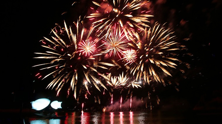 L’image montre un feu d’artifice tiré de nuit au-dessus d’un plan d’eau. Plusieurs gerbes lumineuses de couleur dorée et rouge se déploient dans le ciel sombre. Leur reflet est visible à la surface de l’eau. À gauche de l’image, on distingue des formes lumineuses floues, probablement des installations ou des éclairages proches de la rive. Le ciel est entièrement noir, et aucun autre élément n’est visible en arrière-plan.