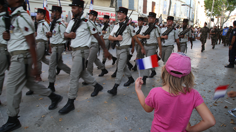 L’image montre un défilé militaire dans une rue. Un groupe de soldats en uniforme marche en formation, portant des bérets noirs, des pantalons clairs, des chemises blanches et des armes sur l’épaule. À l’avant-plan, une enfant de dos, habillée d’un t-shirt rose et d’une casquette rose, tient un petit drapeau tricolore bleu-blanc-rouge. Il y a des spectateurs le long de la rue et des bâtiments en arrière-plan. Le sol est pavé, et on aperçoit des arbres sur la droite de l’image.